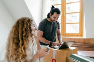 Happy couple unpacking in their new home, using a laptop on a box, sharing coffee and smiles.